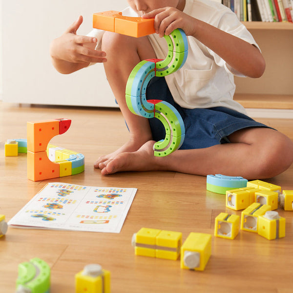 Boy sitting in a playroom and connecting magnetic track blocks following a design from a booklet in front of him.