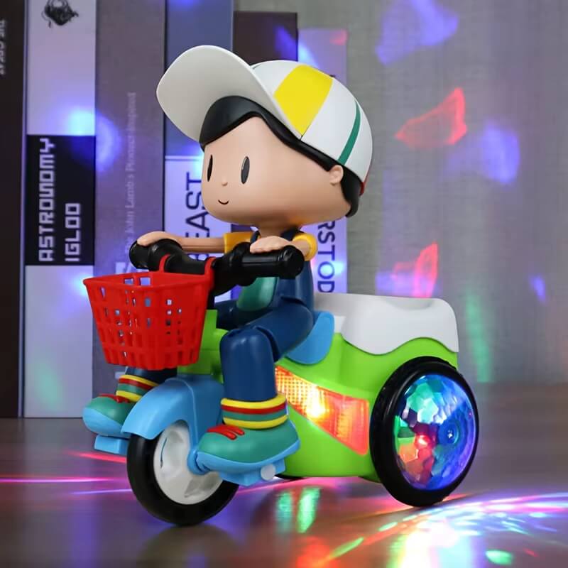 A colorful toy tricycle with a cute cartoon boy figure wearing a cap, sitting on the seat and holding the handlebars. The tricycle features a red front basket, LED lights on the wheels, and a green and blue body. The background has a soft glow with multicolored lights reflecting on the surface.